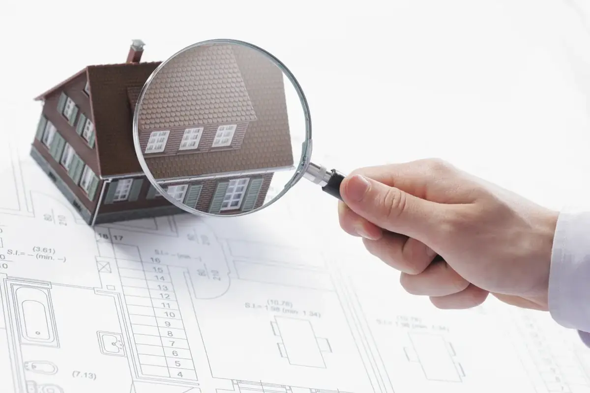 A hand holding a magnifying glass over a model house on a set of building plans